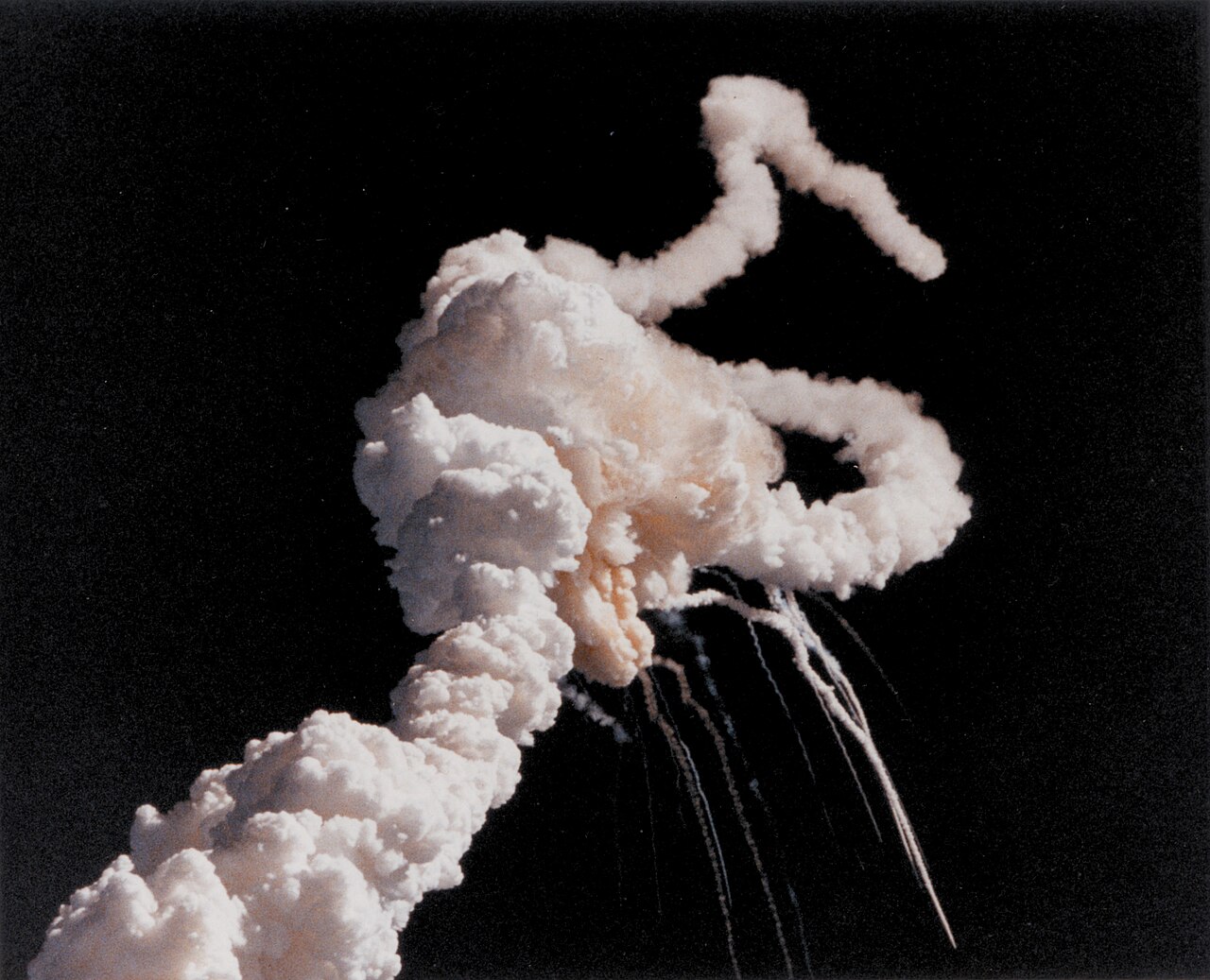Color photograph against a black sky of a large white cloud of smoke with separate plumes curling away from it and trails of debris falling below