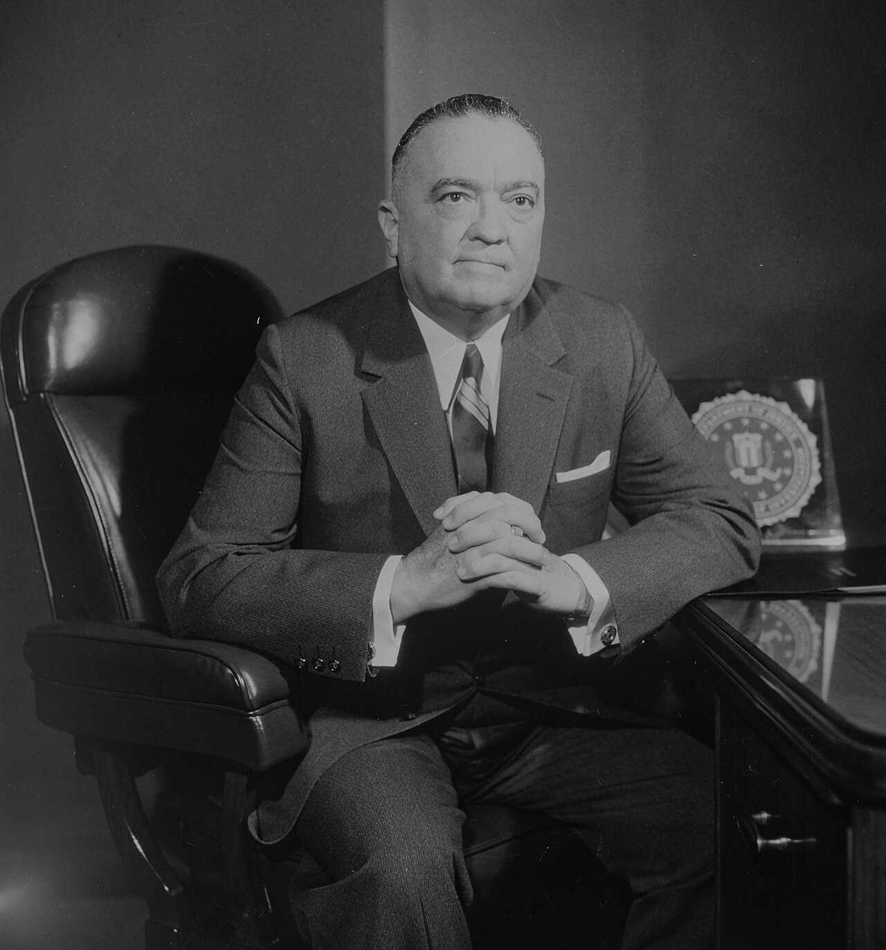 Black and white portrait of a heavyset man in a dark suit and striped tie seated in a leather chair beside a desk, hands clasped, with a plaque bearing the FBI seal on the desk behind him