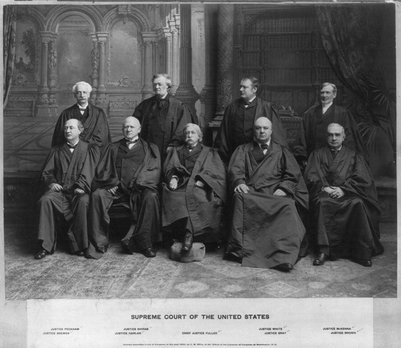 Group photograph of nine Supreme Court justices in black robes, five seated and four standing, in front of a dark backdrop curtain