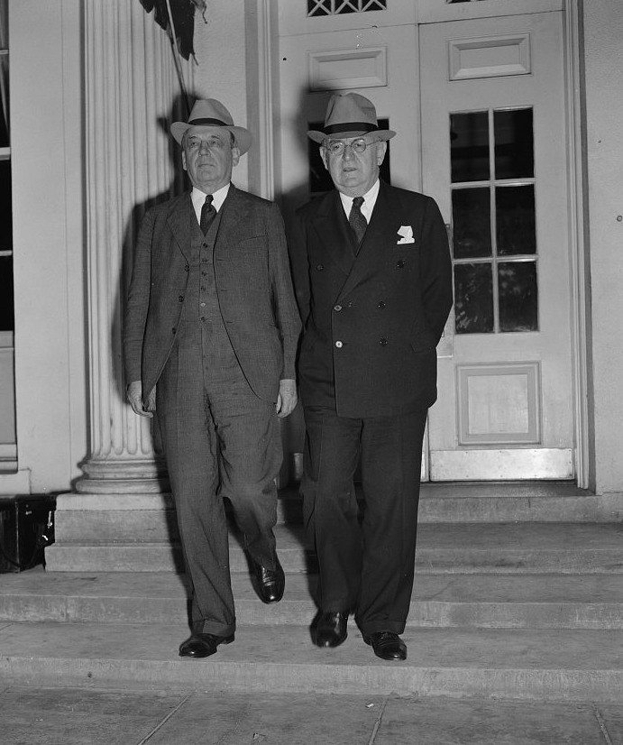 Black and white photograph of two men in suits and hats walking side by side outside a building