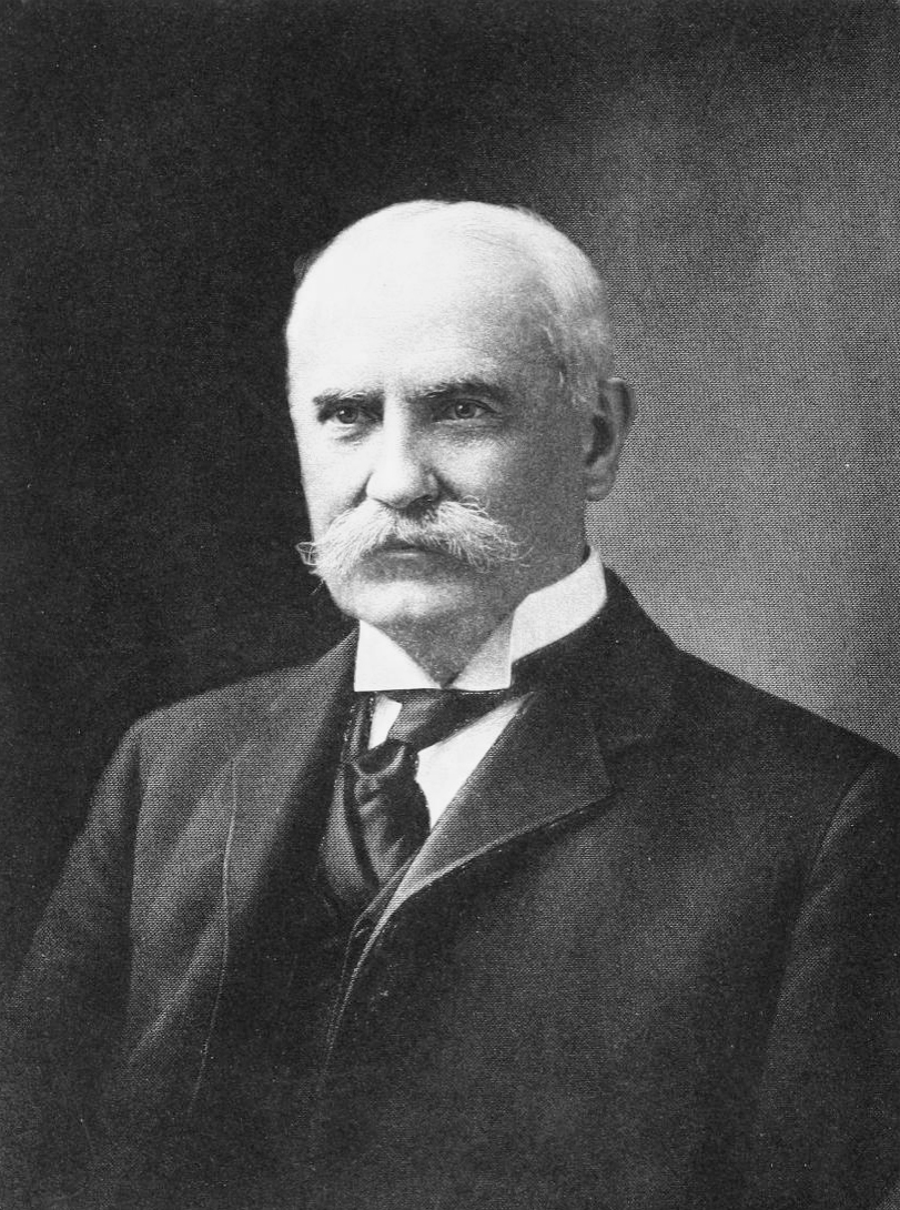 Black and white head and shoulders portrait of an older man with a white mustache in a dark suit and high collar