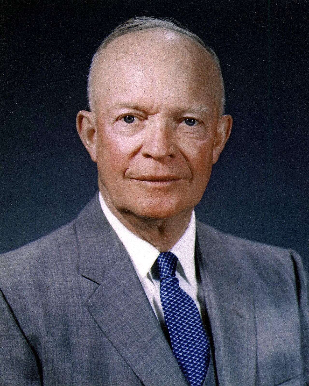 Color official portrait of a bald older man in a dark suit and tie, smiling slightly