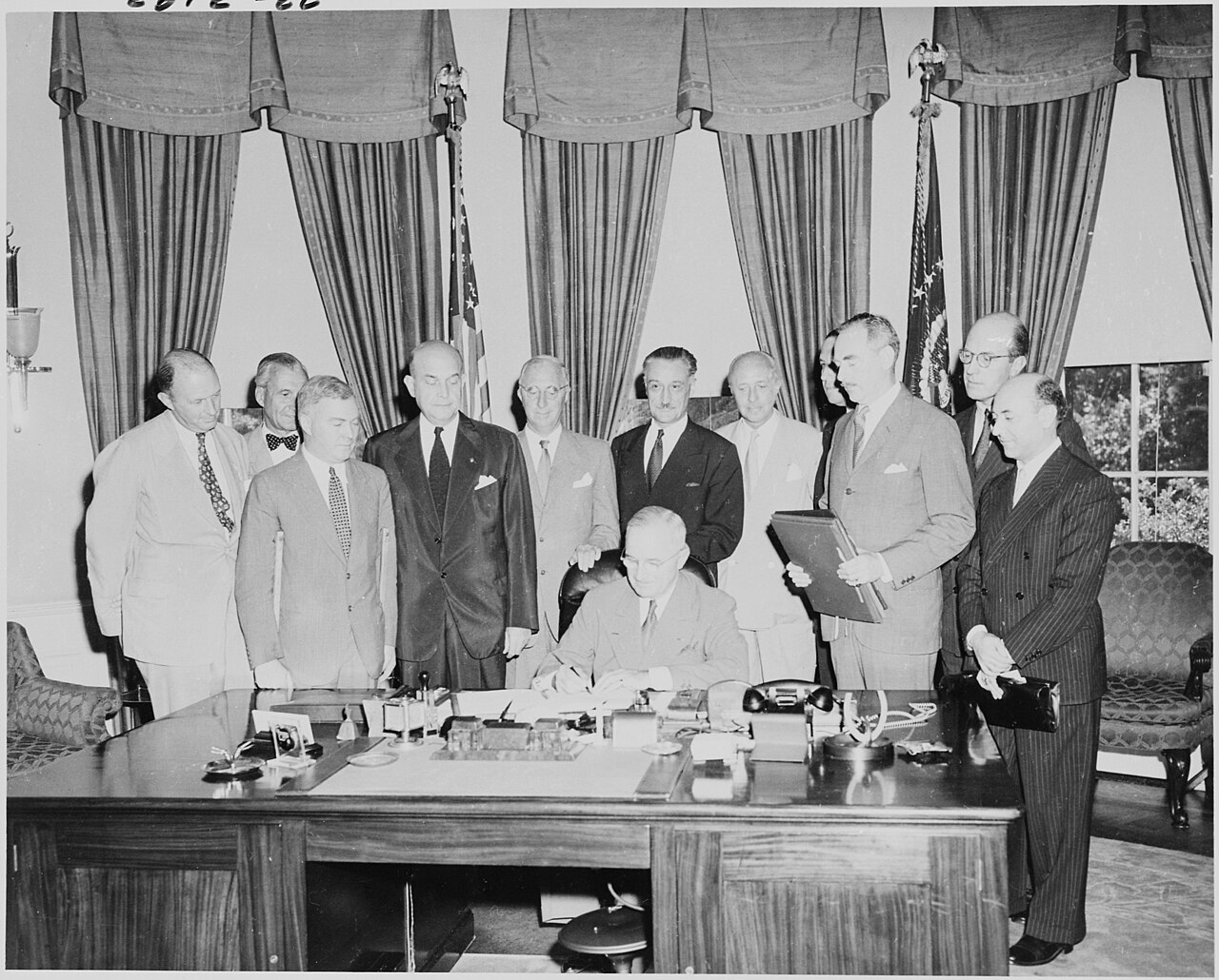 Black and white photograph of President Truman seated at his desk signing a document while diplomats and officials crowd around the desk