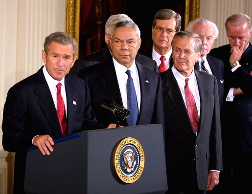 President George W. Bush seated at a desk signing a document, surrounded by standing members of Congress and officials