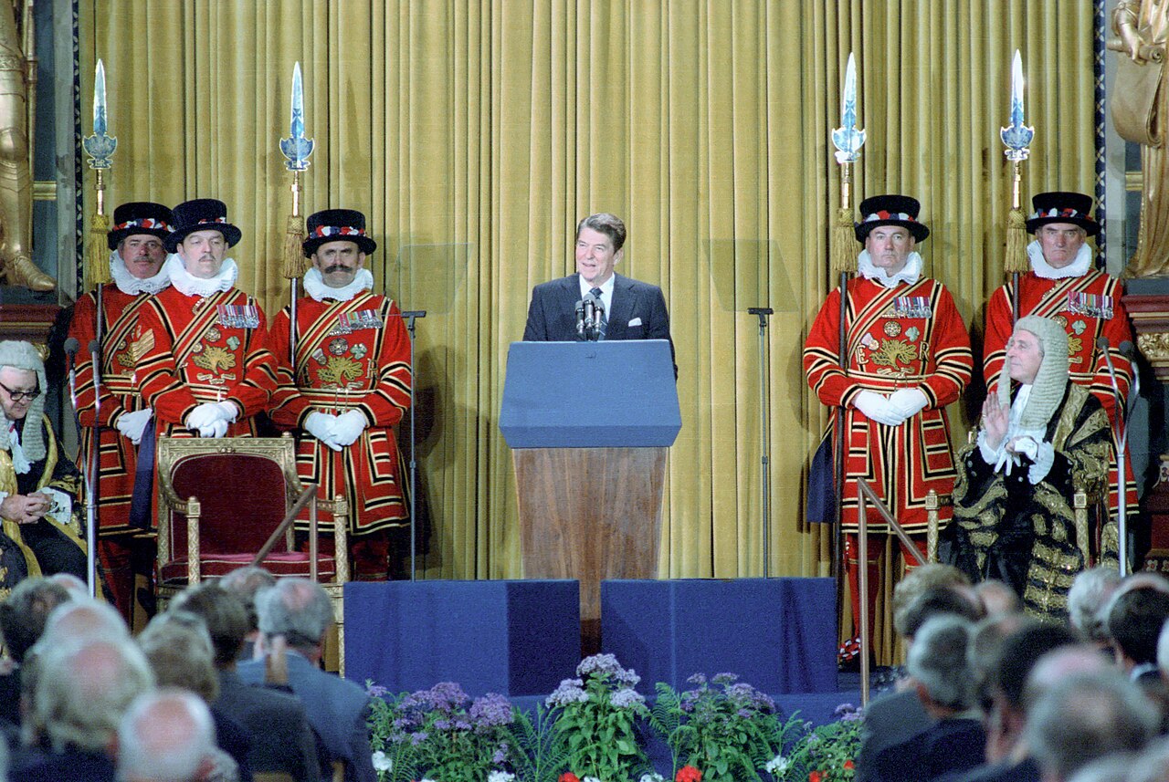 President Reagan standing at a lectern addressing an audience in an ornate hall with tall windows