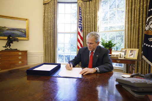 President George W. Bush seated at a desk in the Oval Office signing a document while officials stand behind him