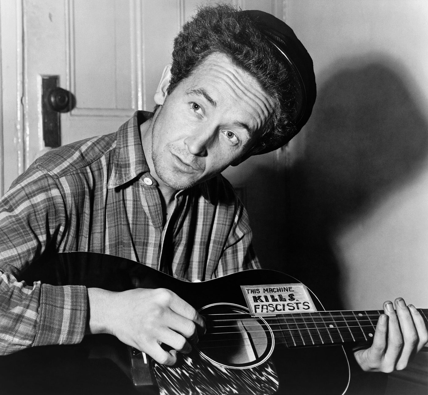 Woody Guthrie seated, playing a guitar with a sticker that reads This Machine Kills Fascists